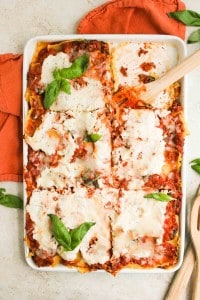 A rectangular baking dish filled with sliced lasagna topped with ricotta cheese, tomato sauce, and basil leaves, with a wooden spatula in one corner.