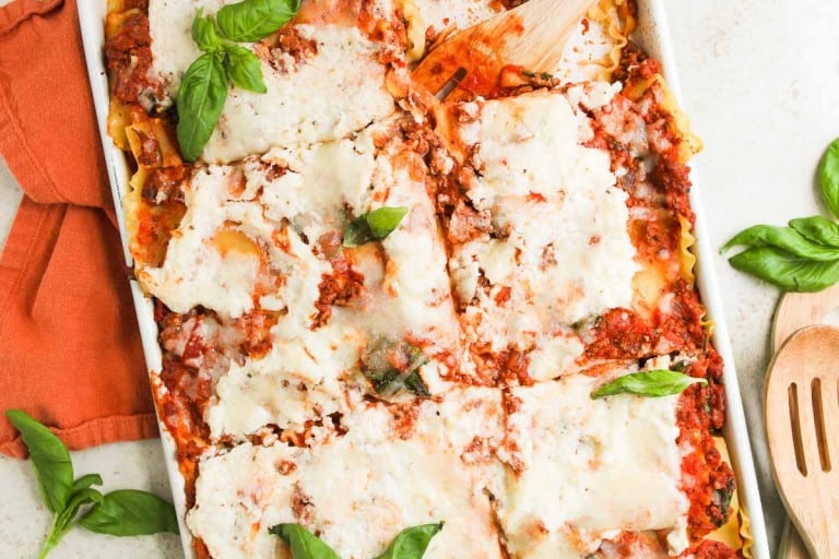 A rectangular baking dish filled with sliced lasagna topped with melted cheese, tomato sauce, ground meat, and basil leaves, with wooden utensils and a red napkin nearby.