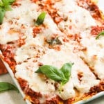 A baked lasagna in a white dish, topped with melted cheese, tomato sauce, and fresh basil leaves, partially sliced and ready to serve.