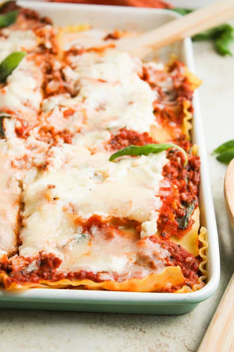 A close-up of a baked lasagna in a rectangular dish, showing layers of pasta, meat sauce, melted cheese, and a few basil leaves.