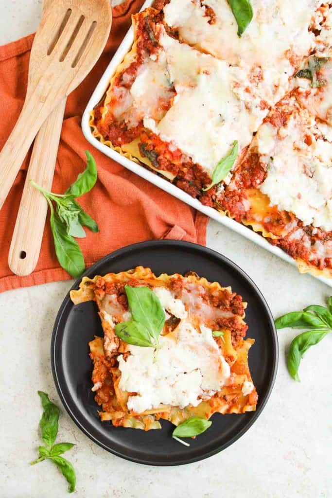 A black plate with two slices of lasagna garnished with basil, next to a baking dish of lasagna, wooden utensils, and a rust-colored napkin.