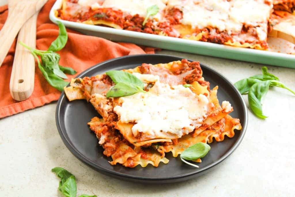 A black plate holds two slices of lasagna topped with melted cheese and fresh basil, with more lasagna and basil leaves in the background.