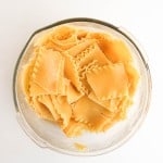 A glass bowl filled with cooked, broken lasagna noodles on a white surface.