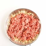 Ground beef and chopped onions cooking in a stainless steel skillet, shown from above on a white background.