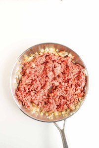 Ground beef and chopped onions cooking in a stainless steel skillet, shown from above on a white background.