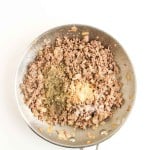 A stainless steel skillet with cooked ground beef, diced onions, and various seasonings on top, viewed from above against a white background.