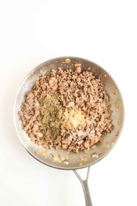 A stainless steel skillet with cooked ground beef, diced onions, and various seasonings on top, viewed from above against a white background.