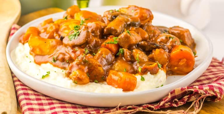 A plate of Slow Cooker Beef Bourguignon with carrots and mushrooms served over mashed potatoes, garnished with chopped herbs, on a checkered cloth.