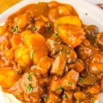 A plate of Slow Cooker Beef Bourguignon stew with potatoes, carrots, and mushrooms in a thick brown sauce served over mashed potatoes, garnished with chopped parsley.
