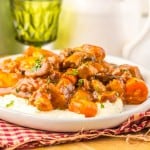 A plate of creamy mashed potatoes topped with hearty Slow Cooker Beef Bourguignon and vegetables, served on a red checkered napkin.