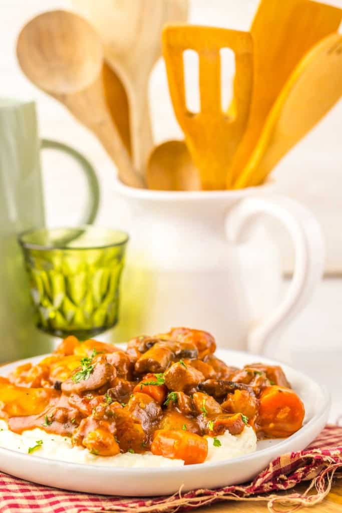 A plate of Slow Cooker Beef Bourguignon with carrots and rich sauce served over mashed potatoes, accompanied by a glass, mug, and wooden utensils in the background.