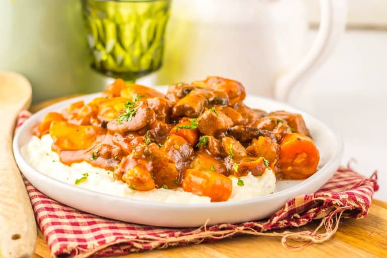 A plate of creamy mashed potatoes topped with hearty Slow Cooker Beef Bourguignon and vegetables, served on a red checkered napkin.