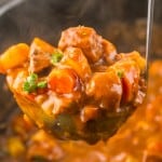 A ladle lifting a serving of Slow Cooker Beef Bourguignon, showcasing chunks of meat, potatoes, carrots, and a parsley garnish.