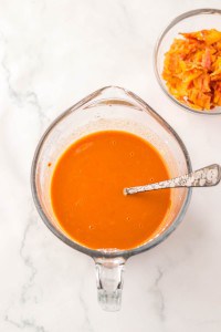 A glass measuring cup filled with orange sauce and a spoon sits next to a small bowl of orange vegetable peels on a white marble surface, perfect for prepping ingredients for Slow Cooker Beef Bourguignon.