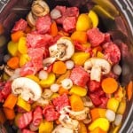 Uncooked stew ingredients for Slow Cooker Beef Bourguignon, including beef chunks, sliced mushrooms, carrots, potatoes, and pearl onions in a slow cooker.