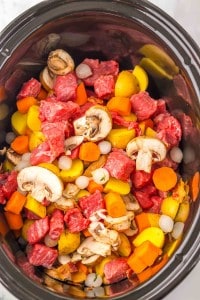 Uncooked stew ingredients for Slow Cooker Beef Bourguignon, including beef chunks, sliced mushrooms, carrots, potatoes, and pearl onions in a slow cooker.