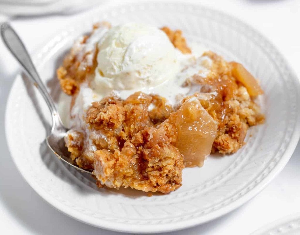A plate of apple crumble topped with vanilla ice cream, with a spoon resting on the side.