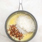 A saucepan containing melted butter, shredded coconut, and chopped pecans—classic ingredients for a German Chocolate Bundt Cake—sits on a light-colored surface.