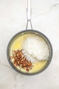 A saucepan containing melted butter, shredded coconut, and chopped pecans—classic ingredients for a German Chocolate Bundt Cake—sits on a light-colored surface.