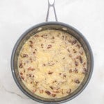 A saucepan filled with a thick, yellowish mixture of chopped pecans and shredded coconut, ready as the signature topping for a German Chocolate Bundt Cake, sits on a white marble surface.