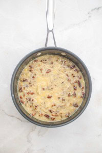 A saucepan filled with a thick, yellowish mixture of chopped pecans and shredded coconut, ready as the signature topping for a German Chocolate Bundt Cake, sits on a white marble surface.