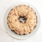 A German Chocolate Bundt Cake topped with a thick layer of coconut and pecan frosting, displayed on a white plate.