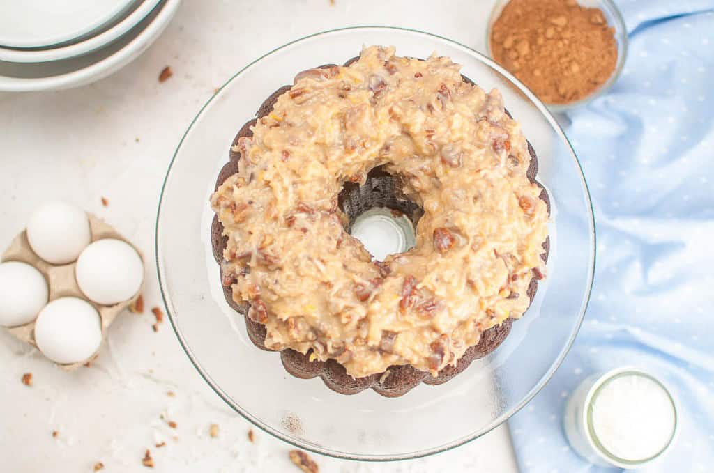 A German Chocolate Bundt Cake topped with coconut pecan frosting sits on a glass cake stand, surrounded by eggs, a bowl of cocoa powder, and a light blue cloth.