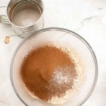 A glass bowl containing sifted flour, cocoa powder, and baking powder sits on a marble surface next to a metal sifter, ready for creating a rich German Chocolate Bundt Cake.
