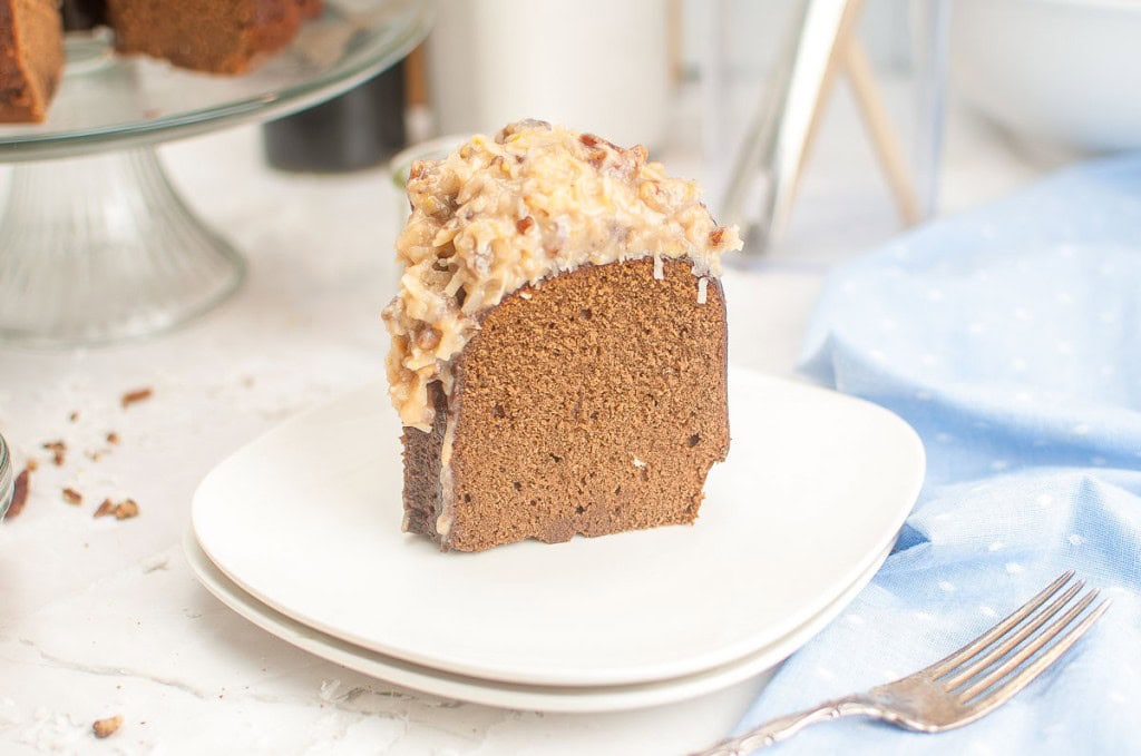 A slice of German Chocolate Bundt Cake with a nutty, coconut topping sits on a white plate next to a fork and a blue napkin.