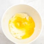 A white mixing bowl containing flour and cracked raw eggs on a light marble surface, ready for creating a delicious German Chocolate Bundt Cake.