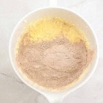 A mixing bowl with yellow batter and cocoa powder partially mixed on top, set on a light marble surface—perfect for starting a rich German Chocolate Bundt Cake.