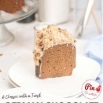 A slice of German Chocolate Bundt Cake with coconut-pecan frosting sits on a white plate; the rest of the decadent cake is visible in the background.