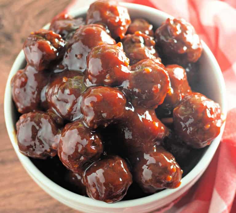 A bowl filled with Slow Cooker Hot Honey Meatballs, glazed in a thick, dark sauce, sits on a wooden surface with a red cloth nearby.