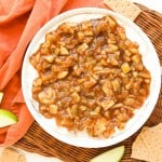 A bowl of apple pie dip topped with spiced apple mixture sits on a wicker tray, surrounded by graham crackers, apple slices, and an orange cloth.