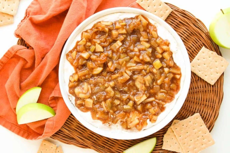 A bowl of apple pie dip topped with spiced apple mixture sits on a wicker tray, surrounded by graham crackers, apple slices, and an orange cloth.