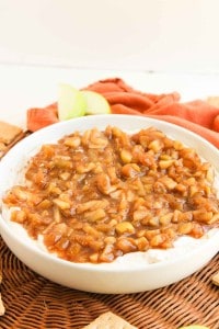 A white bowl filled with cream cheese topped with a chunky apple and cinnamon mixture, placed on a woven mat with apple slices and graham crackers nearby.