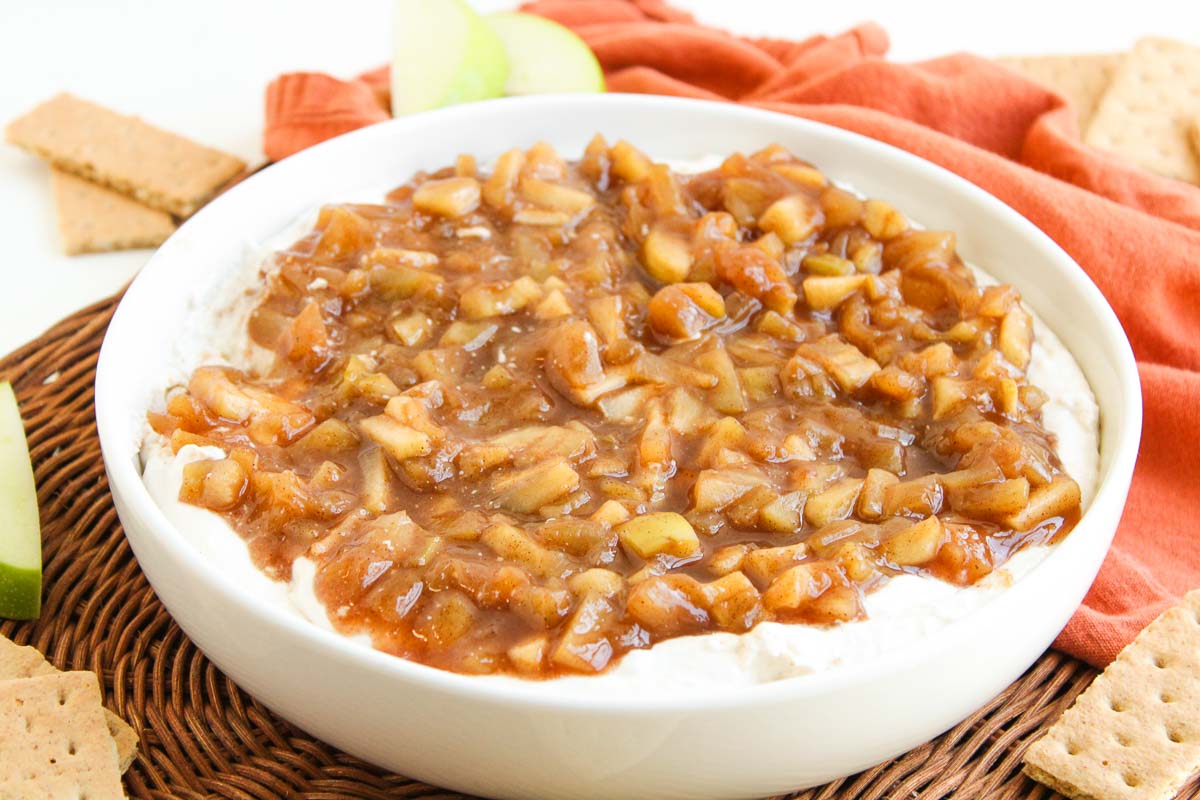 Cream cheese dip topped with warm caramelized apple mixture in a white bowl, surrounded by graham crackers and apple slices.