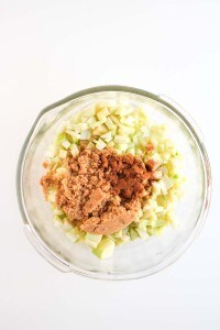 A glass bowl filled with chopped green apples, topped with brown sugar and ground cinnamon, on a white surface.