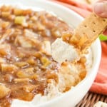 A hand dips a graham cracker into a bowl of cream cheese topped with apple-cinnamon mixture. Crackers are visible around the bowl.