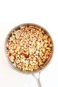 A stainless steel pan with chopped apples and brown liquid, possibly cinnamon and sugar, on a white background.
