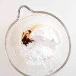 A glass mixing bowl containing whipped topping, powdered sugar, and vanilla extract on a white surface.