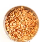 Diced apples coated in a brown sauce are being cooked in a stainless steel pan, viewed from above on a white background.