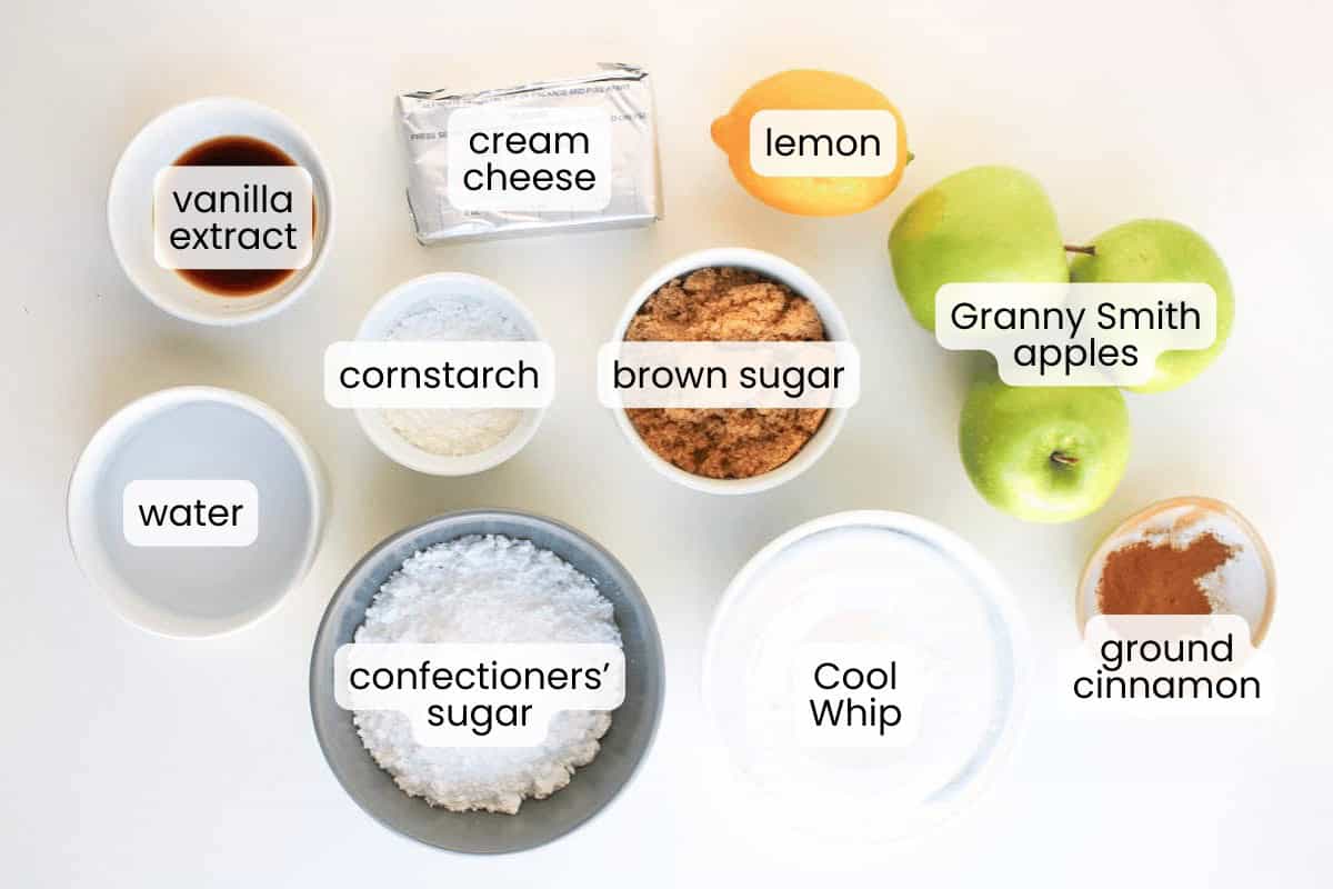Top-down view of labeled baking ingredients for apple pie dip, including cream cheese, lemon, Granny Smith apples, brown sugar, cornstarch, water, vanilla extract, confectioners' sugar, Cool Whip, and ground cinnamon.