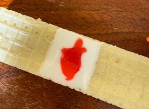 A rectangular wafer with a white cream filling and a small red stain on top sits on a wooden surface.
