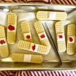 A metal tray holds wafer cookies decorated to look like bandages, with white icing and red jelly, next to a pair of tweezers on a striped cloth.
