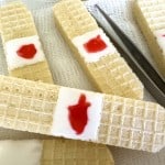 Wafer cookies decorated to resemble bandages, with white icing and red jelly to mimic a wound, placed on gauze with metal tweezers nearby.