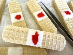 Wafer cookies decorated to resemble bandages, with white icing and red jelly to mimic a wound, placed on gauze with metal tweezers nearby.
