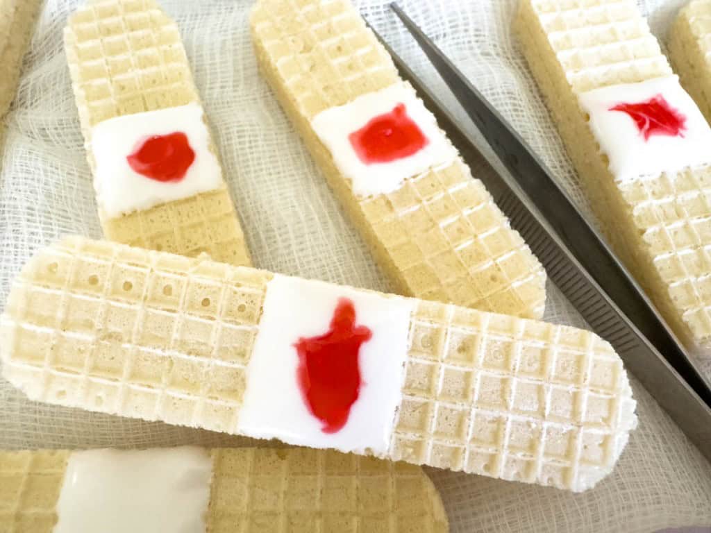 Wafer cookies decorated to resemble bandages, with a white icing "pad" and red jelly "blood," placed on gauze with a pair of tweezers nearby.