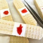 Wafer cookies decorated to resemble bandages, with a white icing "pad" and red jelly "blood," placed on gauze with a pair of tweezers nearby.