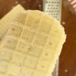 Close-up of a rectangular vanilla wafer cookie held above a brown surface with a metallic grater and red-striped fabric in the background.